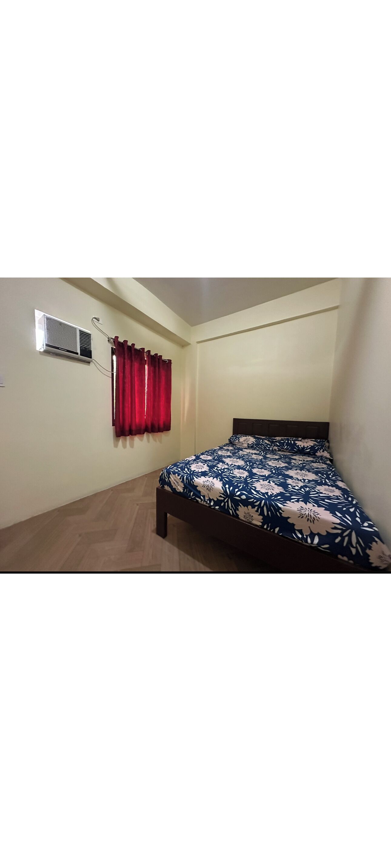A bedroom with a bed, a window with red curtains, and an air conditioning unit on the wall.