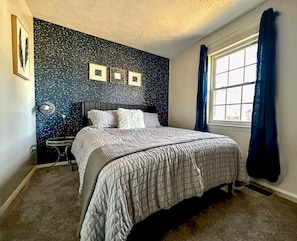 Un dormitorio con cama, mesita de noche, reloj de pared y ventana con cortinas.