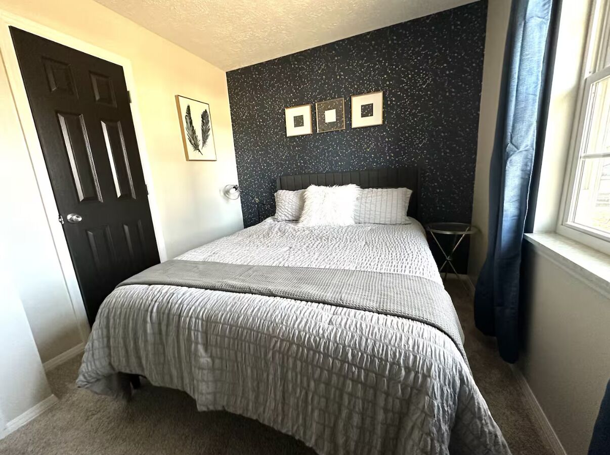 A bedroom with a bed, a door, a wall with three framed pictures, and a window with curtains.