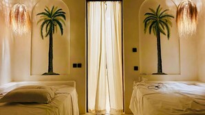 A room with two beds, a ceiling fan, and two palm trees mounted on the walls.