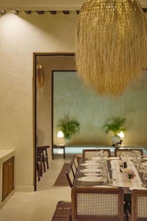 Un espace repas avec une longue table dressée pour le repas, un luminaire suspendu et une vue sur l’extérieur.