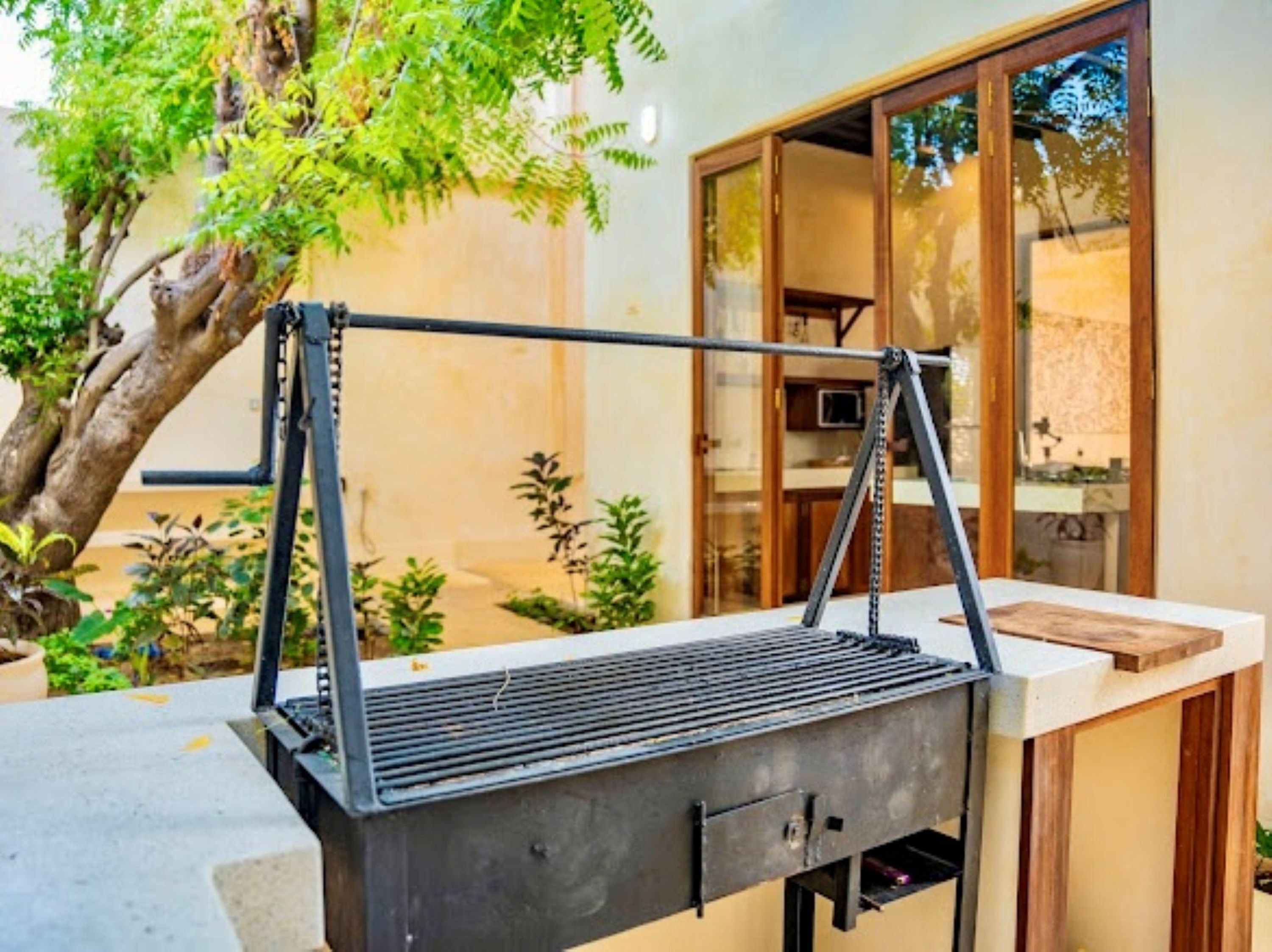Un barbecue d’extérieur doté d’une structure métallique et d’une grille plate, placé sur une terrasse avec un arbre et un bâtiment en arrière-plan.