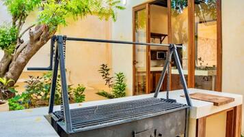 Un barbecue d’extérieur doté d’une structure métallique et d’une grille plate, placé sur une terrasse avec un arbre et un bâtiment en arrière-plan.