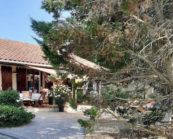 Un edificio con techo de tejas, zona de estar al aire libre con mesas y sillas, y un jardín con diversas plantas y un árbol.