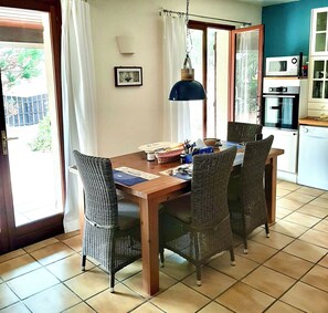 A dining area with wicker chairs, a wooden table, a hanging lamp, and a kitchen with appliances. - Comfortable vacation house a few steps from the ocean in Hourtin-Plage (Hourtin)