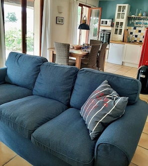 A blue sofa with a striped pillow in a room with a dining table, chairs, and a kitchen area.