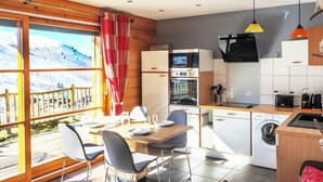 A compact kitchen with a microwave, oven, and washing machine, a dining area with a table and chairs, and a large window with a view of a snowy landscape. - Chalet apartment for rent with south-facing terrace and private sauna (saint francois longchamp)
