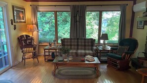 Una sala de estar con un sofá de cuadros, una mesa de centro de madera, un sillón mecedora y vistas al verdor a través de grandes ventanales.