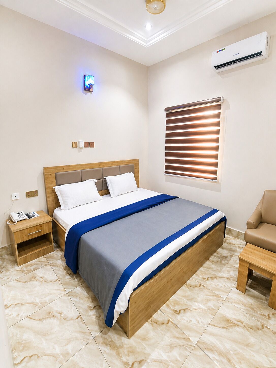 A hotel room with a bed, bedside table, lamp, air conditioning unit, window with blinds, and a small wooden table.