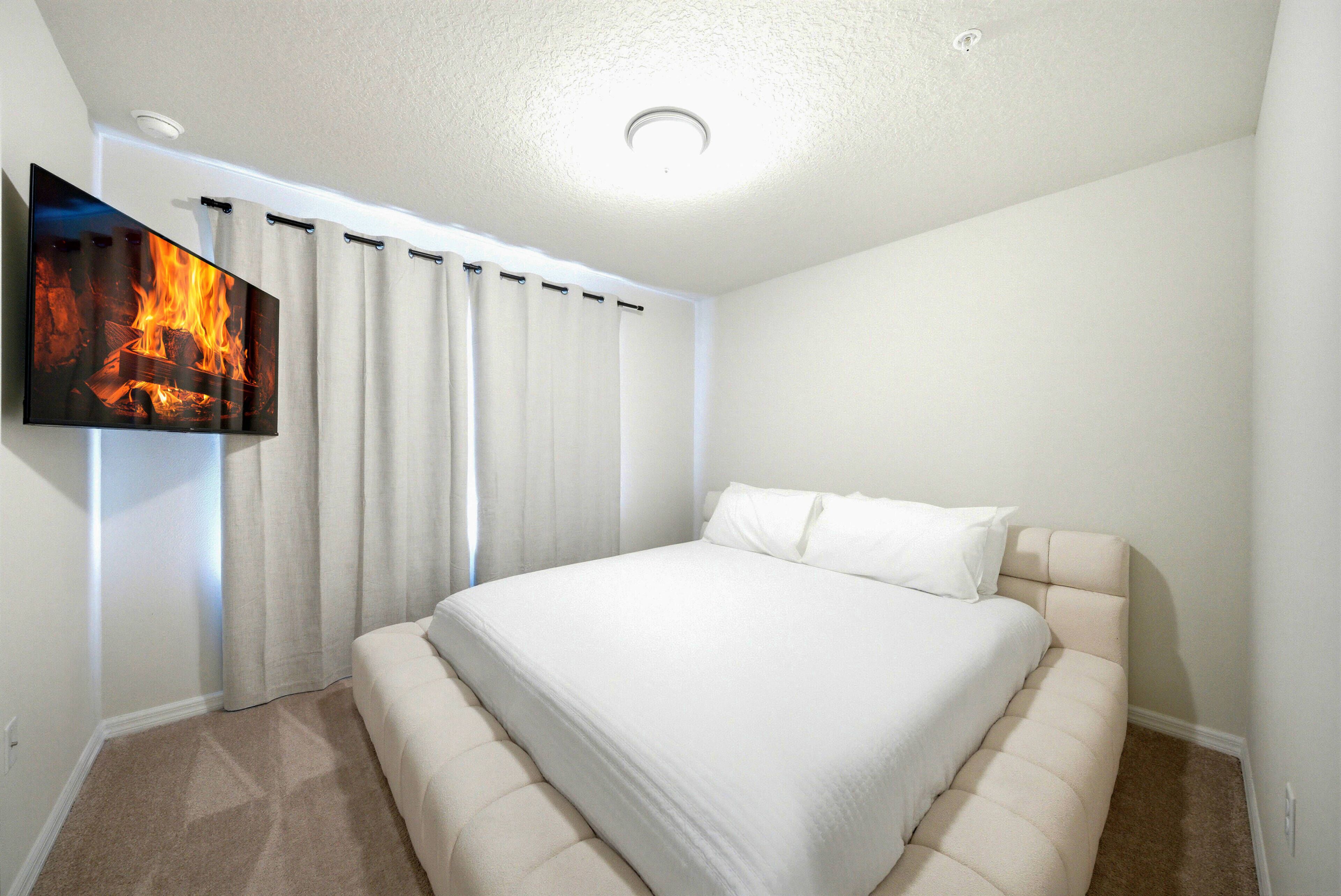 This cozy bedroom invites relaxation with its plush light-colored bed and soft beige curtains framing the window. A sleek flat-screen TV adds entertainment, enhancing your stay in this tranquil haven after a day of adventure.