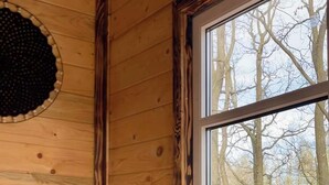 Ein Interieur einer Holzhütte mit Holzwänden, einem grauen Sofa, einem runden Tisch und einem großen Fenster mit Blick auf Bäume.
