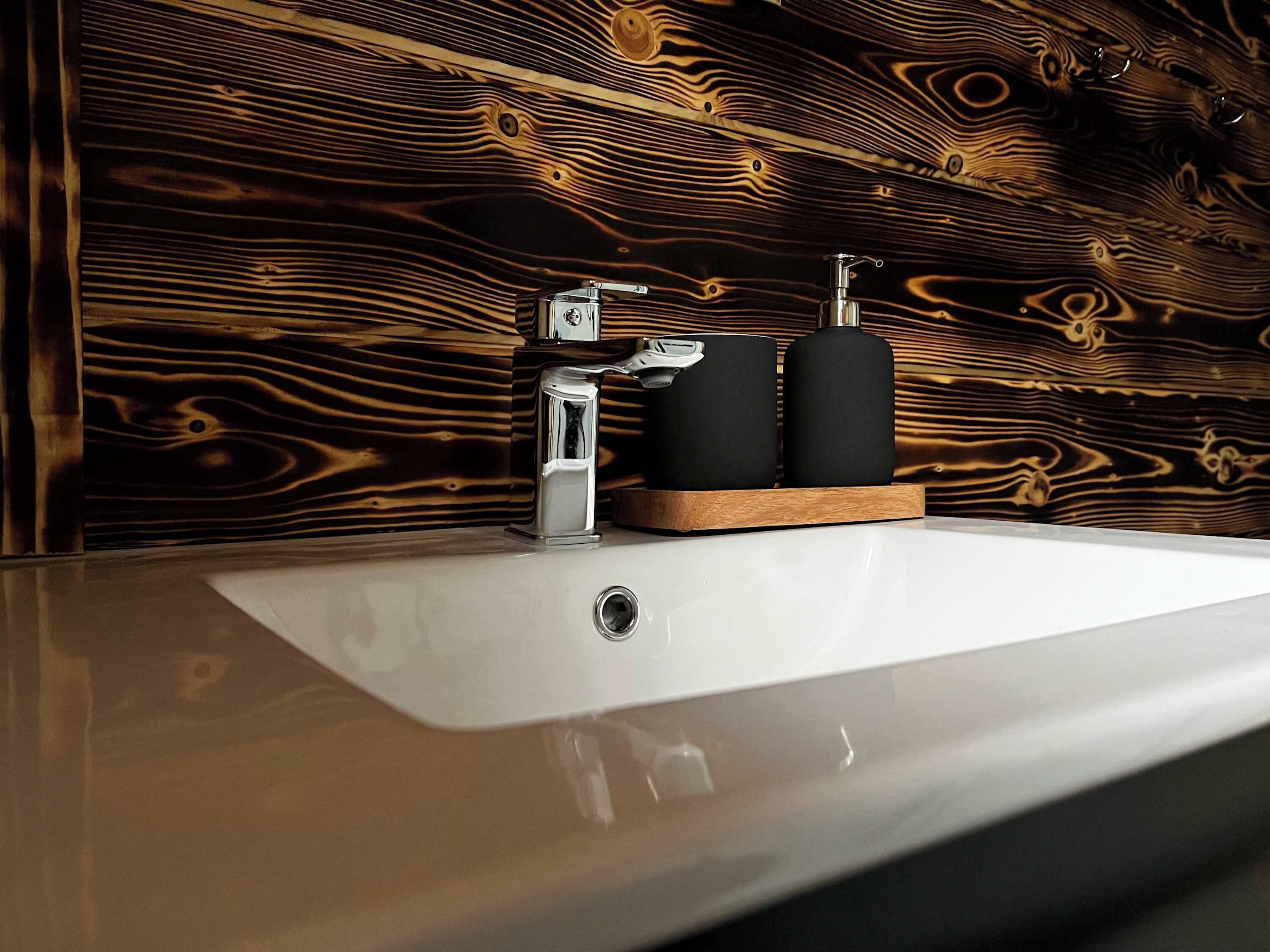 Un baño con pared de madera, un lavamanos blanco, un grifo cromado y dos recipientes negros cilíndricos sobre una bandeja de madera.