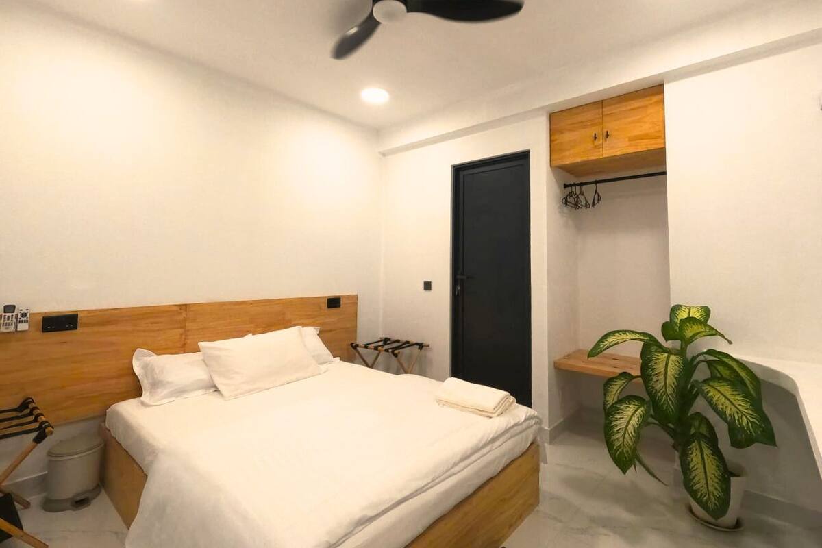 Quarto com cama, ventoinha no teto, porta escura, armário de madeira e planta em vaso.