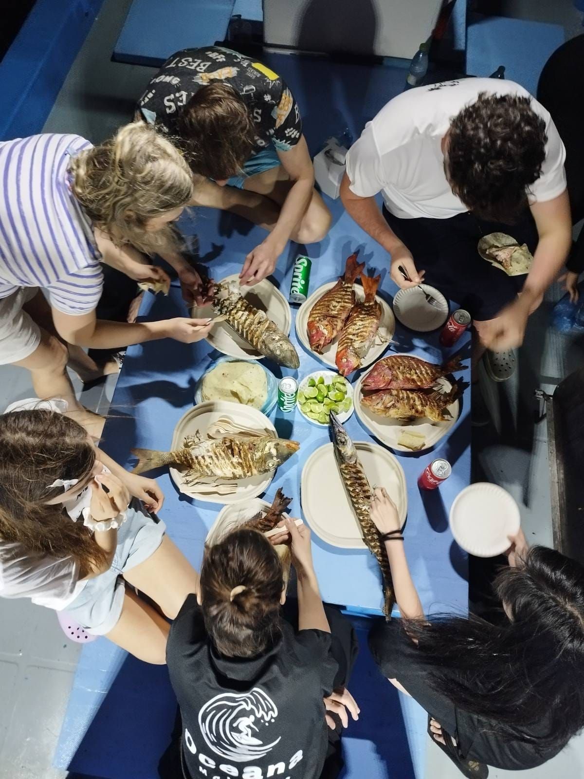 A group of people around a table with various dishes, including grilled fish and a can of soda.