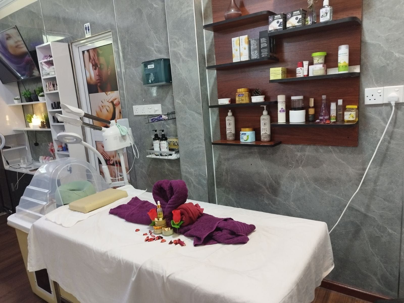 Un cuarto de tratamientos de spa con una camilla de masaje, un estante empotrado con diversos productos para el cuidado de la piel y un cartel en la pared.