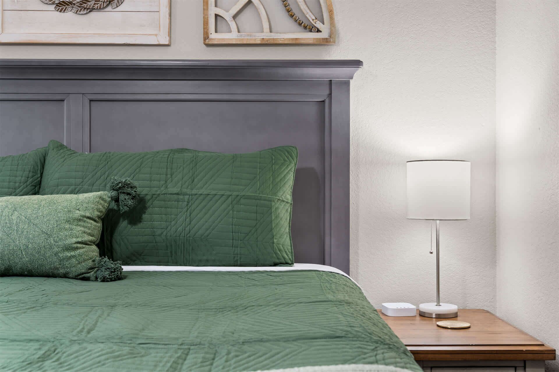 Una cama bien hecha con ropa de cama verde, una lámpara de noche y una mesita de noche pequeña con una lámpara y un dispositivo.