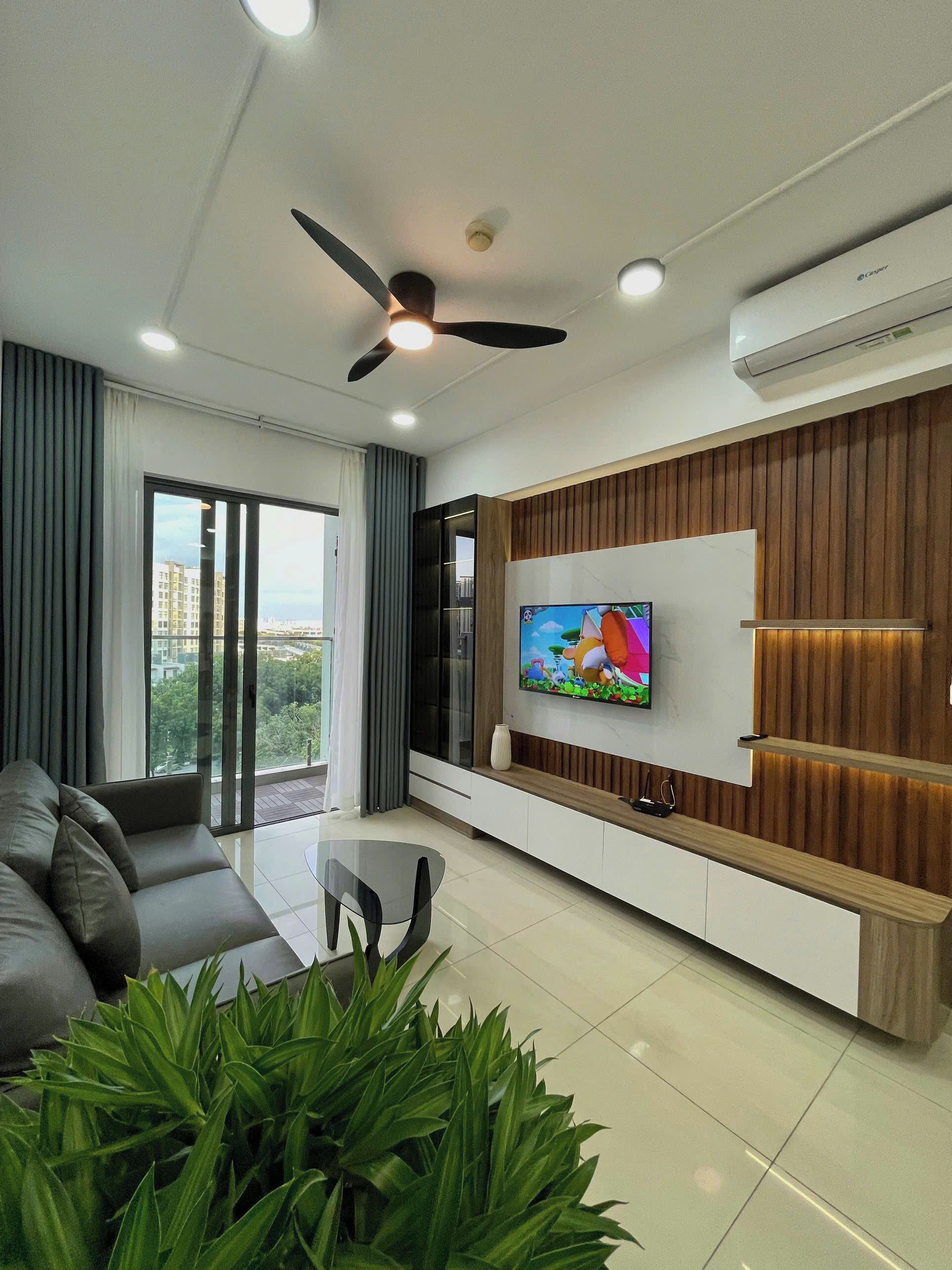 A modern living room with a flat-screen TV, a ceiling fan, and a large window.