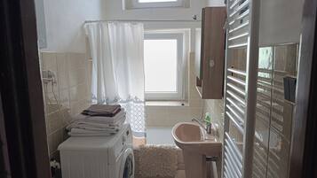 A compact bathroom with a washing machine, a sink, a towel rack, and a window with a curtain.
