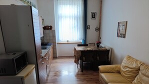 Cocina compacta con refrigerador, microondas y gabinetes de madera. Un comedor con sillas está ubicado cerca de una ventana con cortinas. Un sofá de cuero se encuentra en la sala.