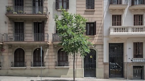 Un immeuble à plusieurs étages, avec un arbre devant, des balcons dotés de garde-corps en métal et une porte avec une plaque numérotée.