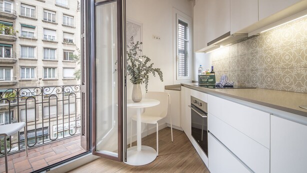 Une cuisine moderne dotée d’une grande fenêtre, d’un coin repas blanc et offrant une vue sur un immeuble avec des balcons.