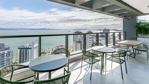 Un balcon avec vue sur la mer et les bâtiments. On y trouve des tables et des chaises pour prendre ses repas.