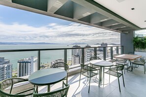 Varanda com vista para o mar e edifícios, equipada com mesas e cadeiras.