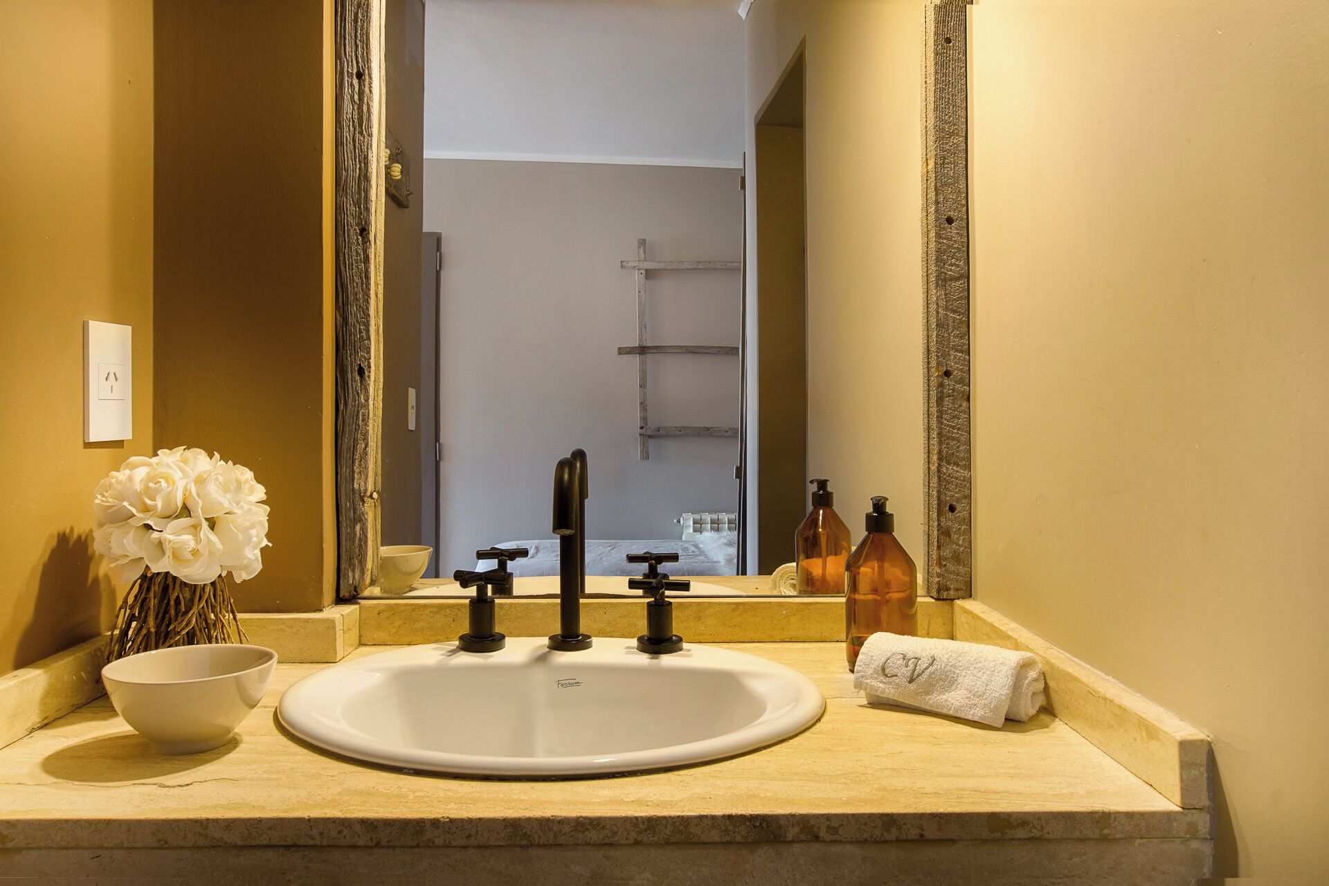 Un bagno con un lavandino bianco, due rubinetti neri, uno specchio, un asciugamano bianco e due flaconi ambrati.