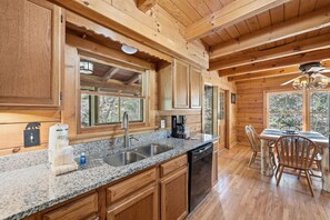A wooden cabin interior with a kitchen featuring granite countertops, a dishwasher, and a dining area with wooden chairs and a table. - Be Still | MTN Views, Hot Tub, Grill, Fireplace! (Sevierville)