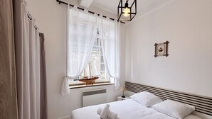 Une chambre à coucher avec un grand lit, une fenêtre ornée de rideaux légers, une petite table sur laquelle est posé un modèle de bateau, une lampe de chevet et un tableau encadré accroché au mur.