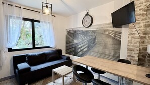 Une pièce moderne avec un canapé noir, une petite table en bois, un coin repas avec des chaises, une grande horloge et un téléviseur à écran plat fixé à un mur en pierre.
