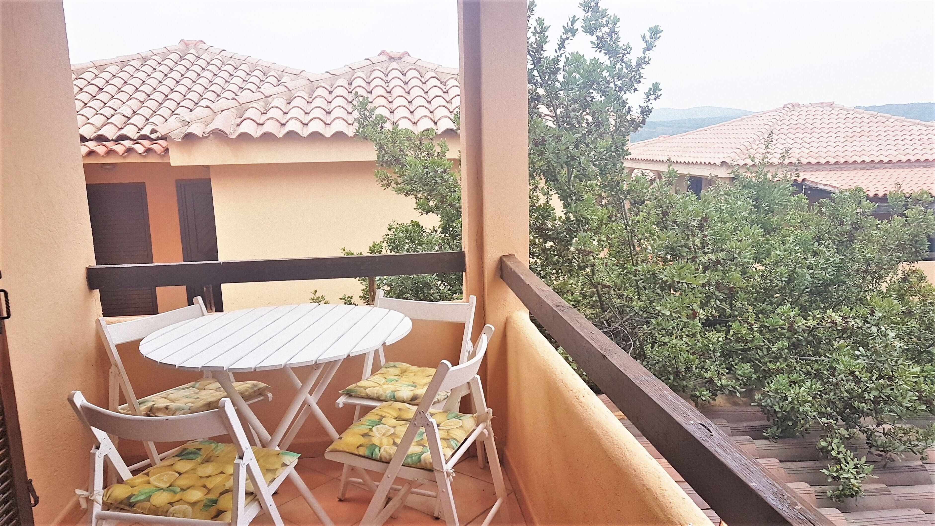 A balcony with a white table and chairs, a view of neighboring buildings with terracotta roofs, and a wooden railing.