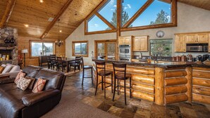A log cabin interior with a stone fireplace, wooden ceiling, large windows, a kitchen with wooden cabinets, and a bar area.