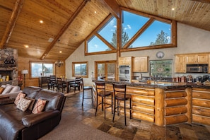 A log cabin interior with a stone fireplace, wooden ceiling, large windows, a kitchen with wooden cabinets, and a bar area.