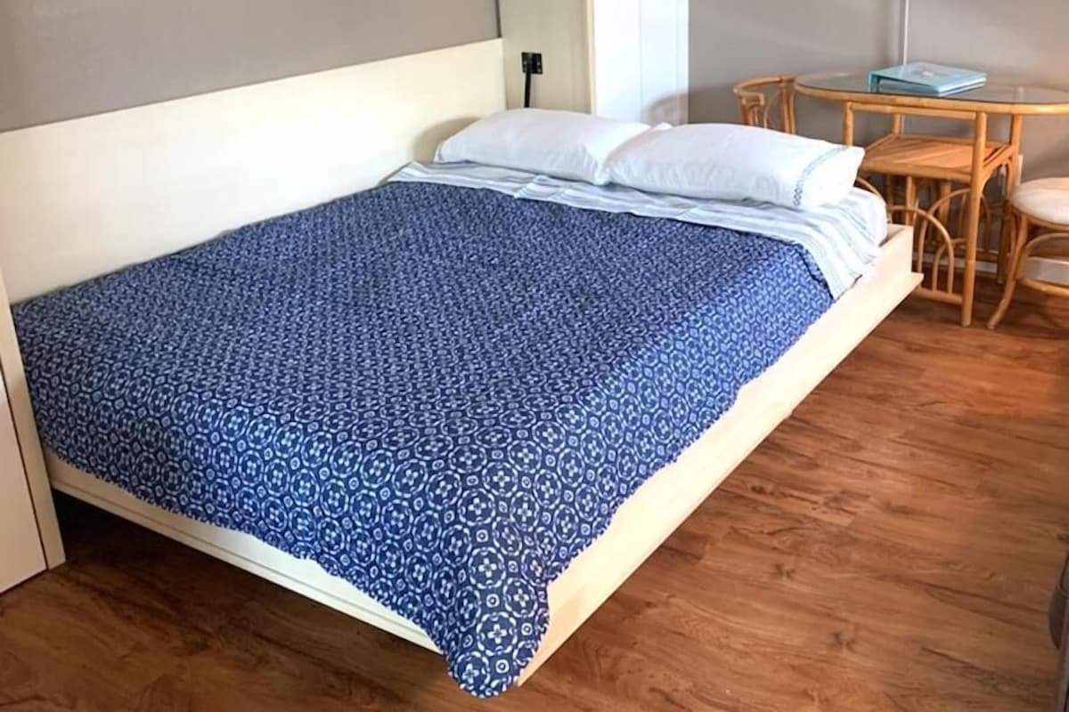 A bed with a patterned bedspread, a bedside table, and a chair in a room with wooden flooring and a painting on the wall.