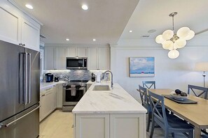 A modern kitchen with stainless steel appliances, a marble island, and a dining area with a view of the beach. - Kiawah Exclusives | 5117 Windswept Villa (Kiawah Island)