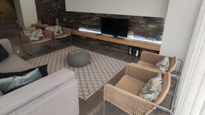 A modern living room with a stone wall, a flat-screen TV, wicker furniture, and a patterned rug. - Local Accommodation – Douro Barcos House (Barcos)