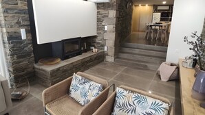 A living room with a fireplace, stone walls, wicker furniture, and a kitchen in the background. - Local Accommodation – Douro Barcos House (Barcos)