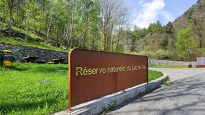 Opastekyltti, jossa lukee ”Réserve naturelle du lac de Villard”, taustalla kivimuuri ja piknikpöytiä.