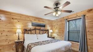 Une chambre à coucher aux murs lambrissés, avec un lit, deux lampes de chevet, un ventilateur de plafond et une fenêtre avec des rideaux.