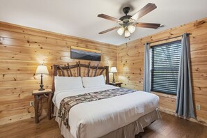 A wooden-paneled bedroom with a bed, two bedside lamps, a ceiling fan, and a window with curtains. - Memory Maker Lodge:Hot Tub, Multicade, Grill, MORE (Gatlinburg)
