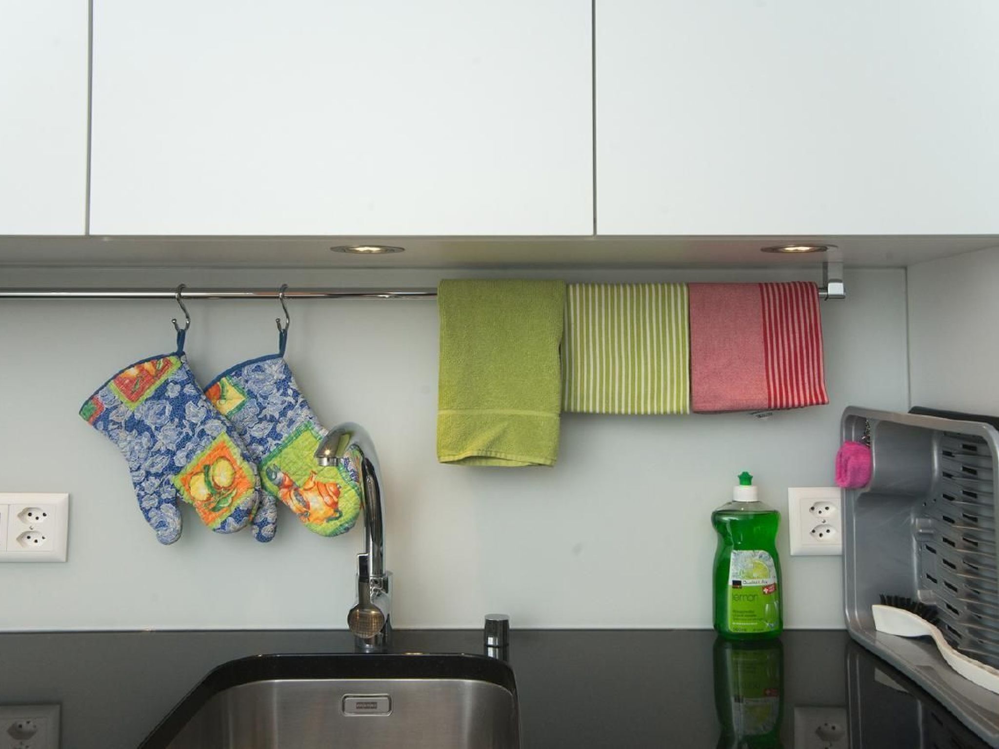 A kitchen with a sink, a faucet, and a dish rack. There are two colorful oven mitts hanging on the wall.