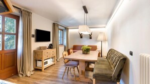 Ein gemütliches Wohnzimmer mit einem Holzesstisch, einer Couch, einem an der Wand montierten Fernseher und einem Fenster mit Vorhängen.