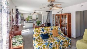 A living room with a sofa, a dining table, chairs, a ceiling fan, a cabinet, and a kitchen in the background.