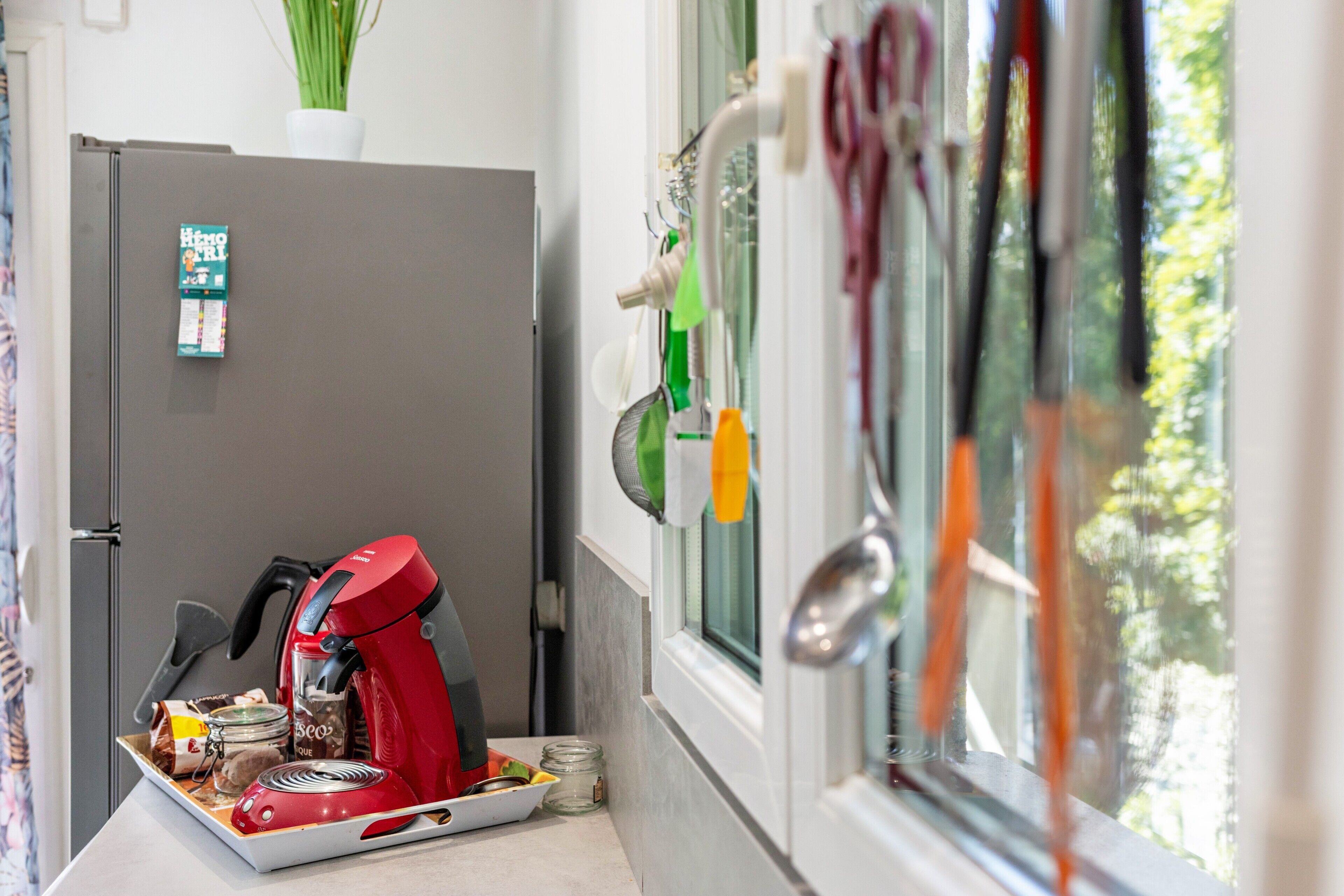Una cocina con un refrigerador, una cafetera roja y una ventana que da a vistas de vegetación.