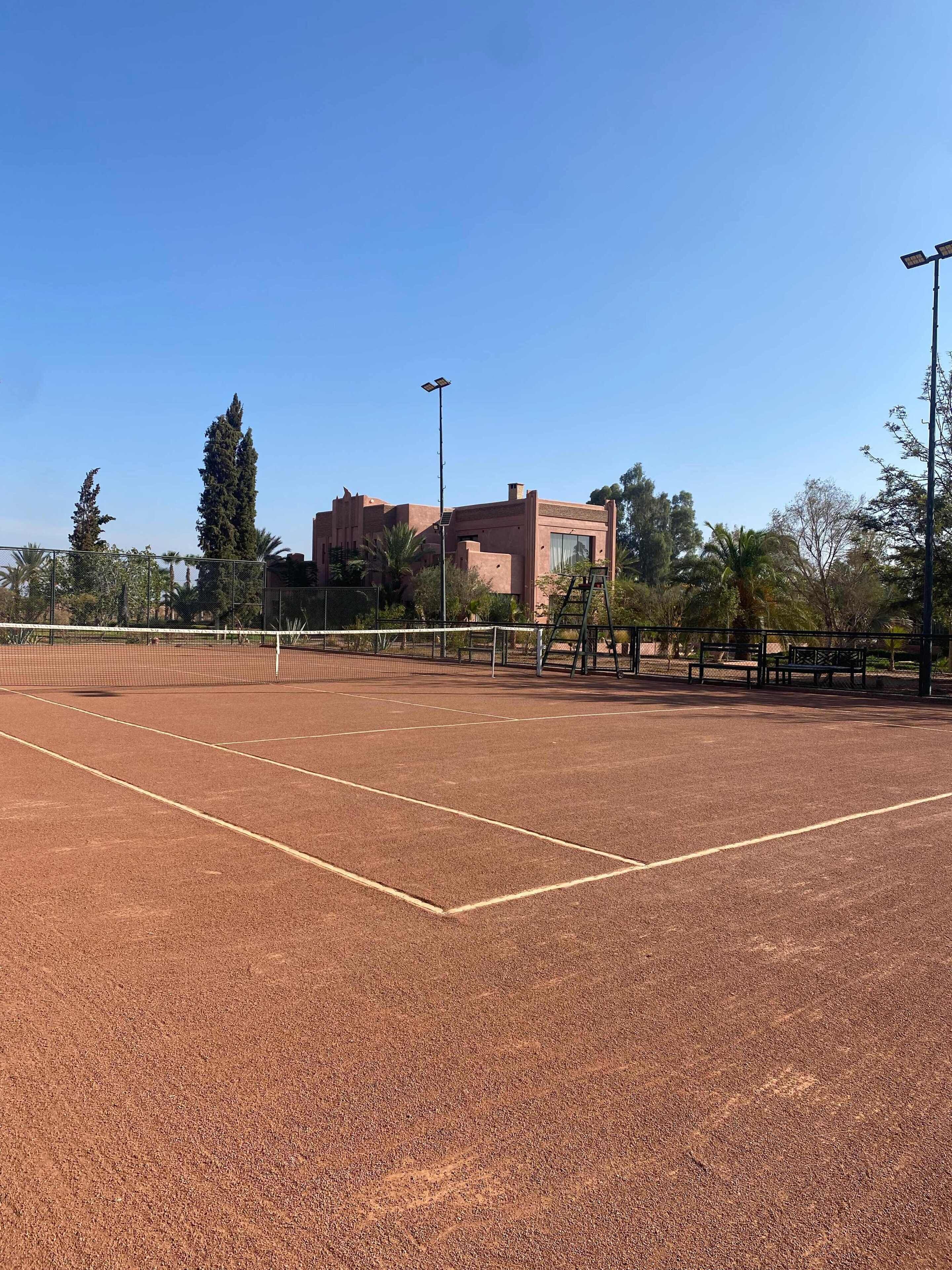 Een buiten tennisbaan met een net, omringd door een hek, en een gebouw op de achtergrond.