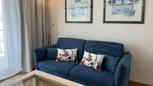 A blue sofa with floral cushions, a glass coffee table, and two framed artworks on the wall. - Chesa Quadrella by Interhome (Pontresina)