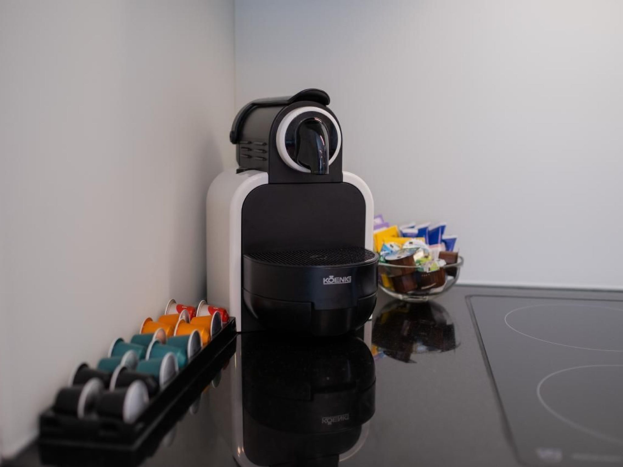 A coffee machine with a control panel, a tray of coffee pods, and a bowl of snacks on a reflective surface.