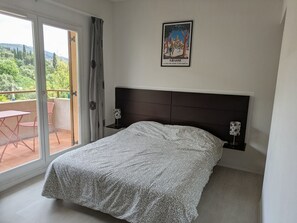 A bedroom with a bed, a nightstand, a framed picture, and a balcony with a view of greenery. - Charming studio for 2 people, near the shops (Grasse)