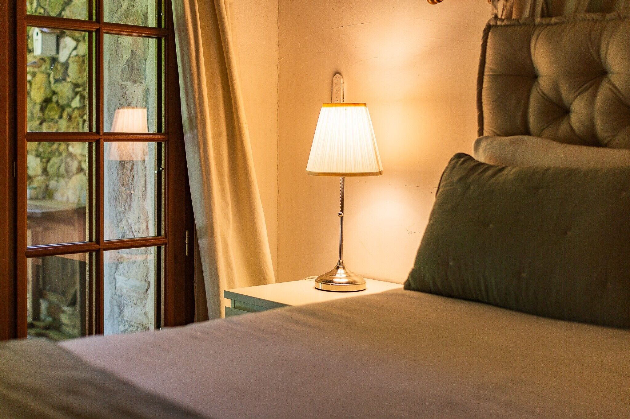 Habitación de hotel con una cama, una mesita de noche con lámpara, una ventana con cortinas y vistas a zonas verdes.